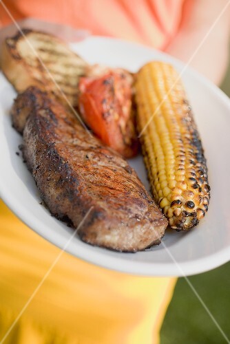Frau hält Teller mit gegrilltem Rindersteak und Maiskolben
