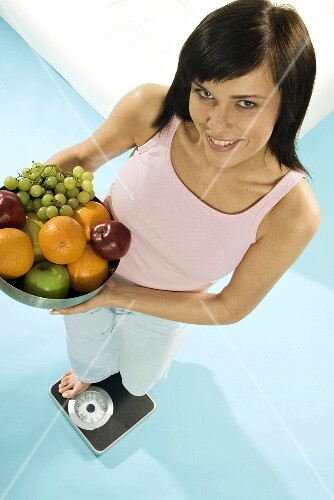 Young woman standing on scales with bowl of fruit
