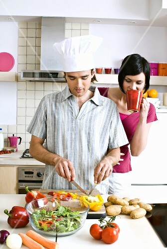Man preparing salad, woman with mug of coffee watching