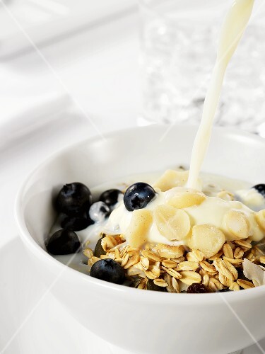 Pouring milk on to muesli with yoghurt, blueberries & nuts