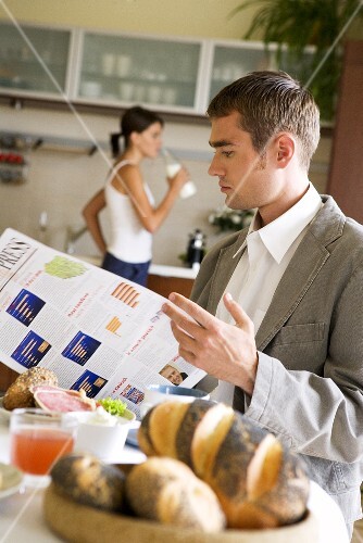 Man reading newspaper at breakfast table, woman drinking milk